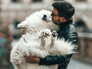 メガネをかけて髭を蓄えた男性が白くて大きな犬を両手で抱き抱えている。仰向けに丸まった犬は気持ちよさそうな表情